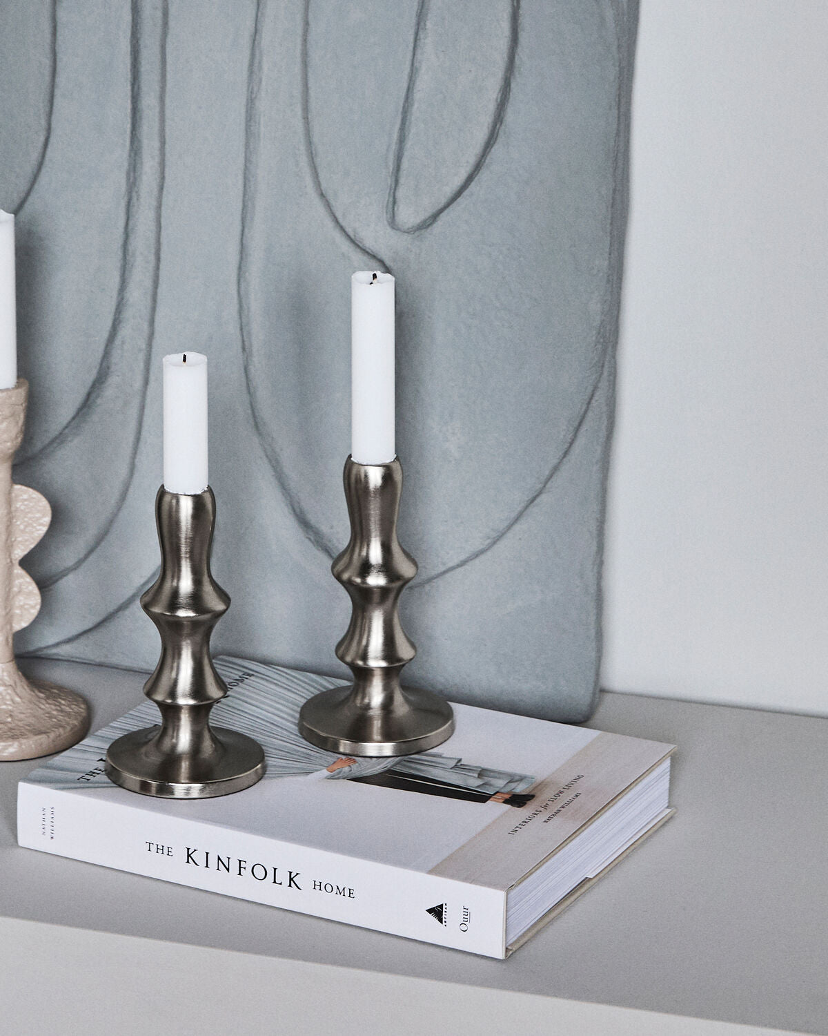 House Doctor Reese candle stands in Silver Finish, styled with lit candles on a book next to another candle holder