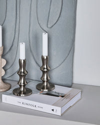 House Doctor Reese candle stands in Silver Finish, styled with lit candles on a book next to another candle holder