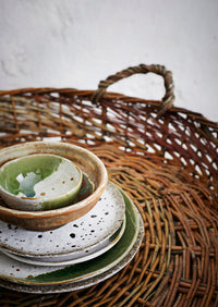 Stack of ceramic plates and bowls with a white green finish on a woven basket with a neutral background