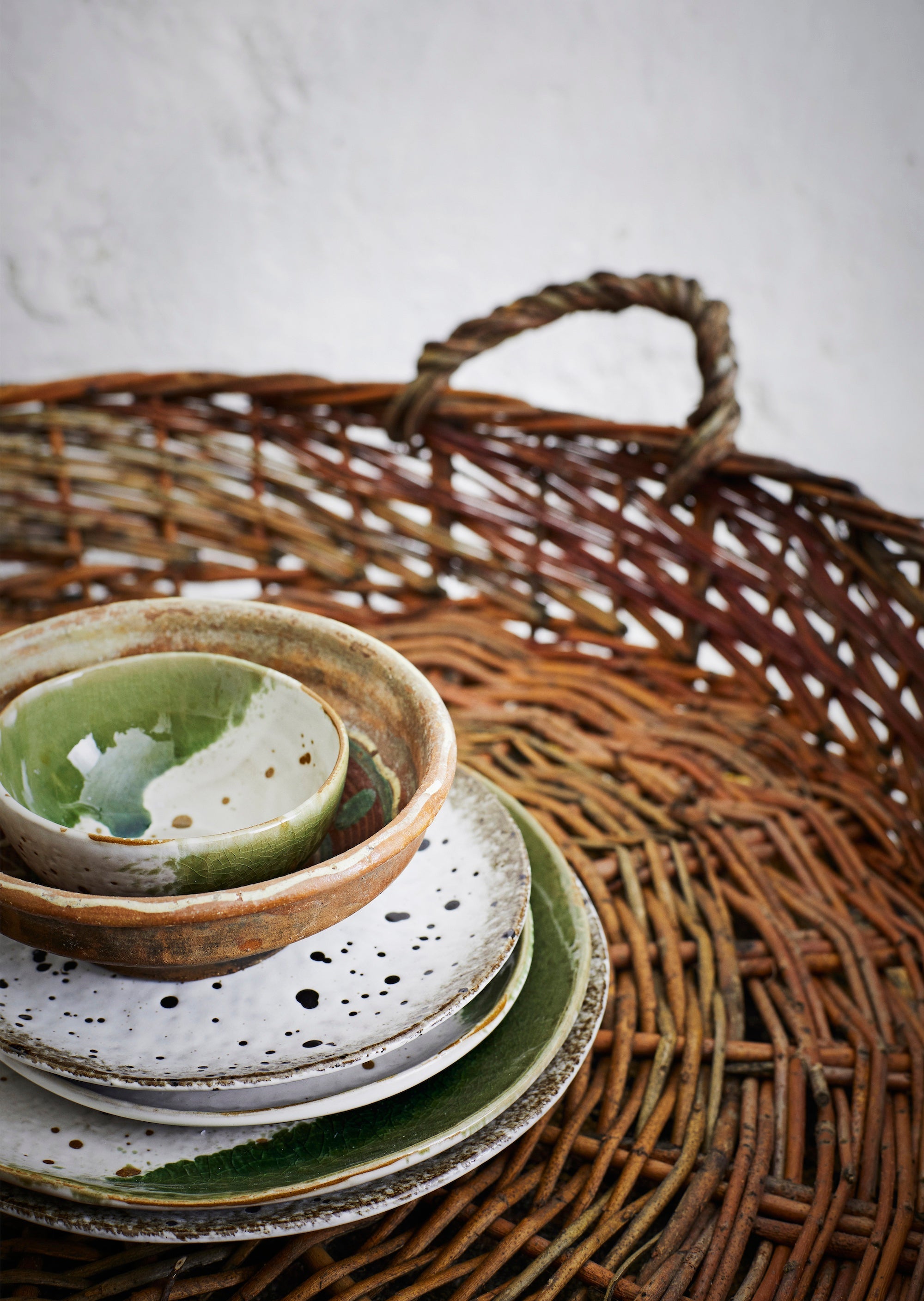 Stack of ceramic plates and bowls with a white green finish on a woven basket with a neutral background