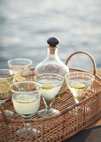 Cocktail glasses with lemon slices on a wicker tray by a body of water.