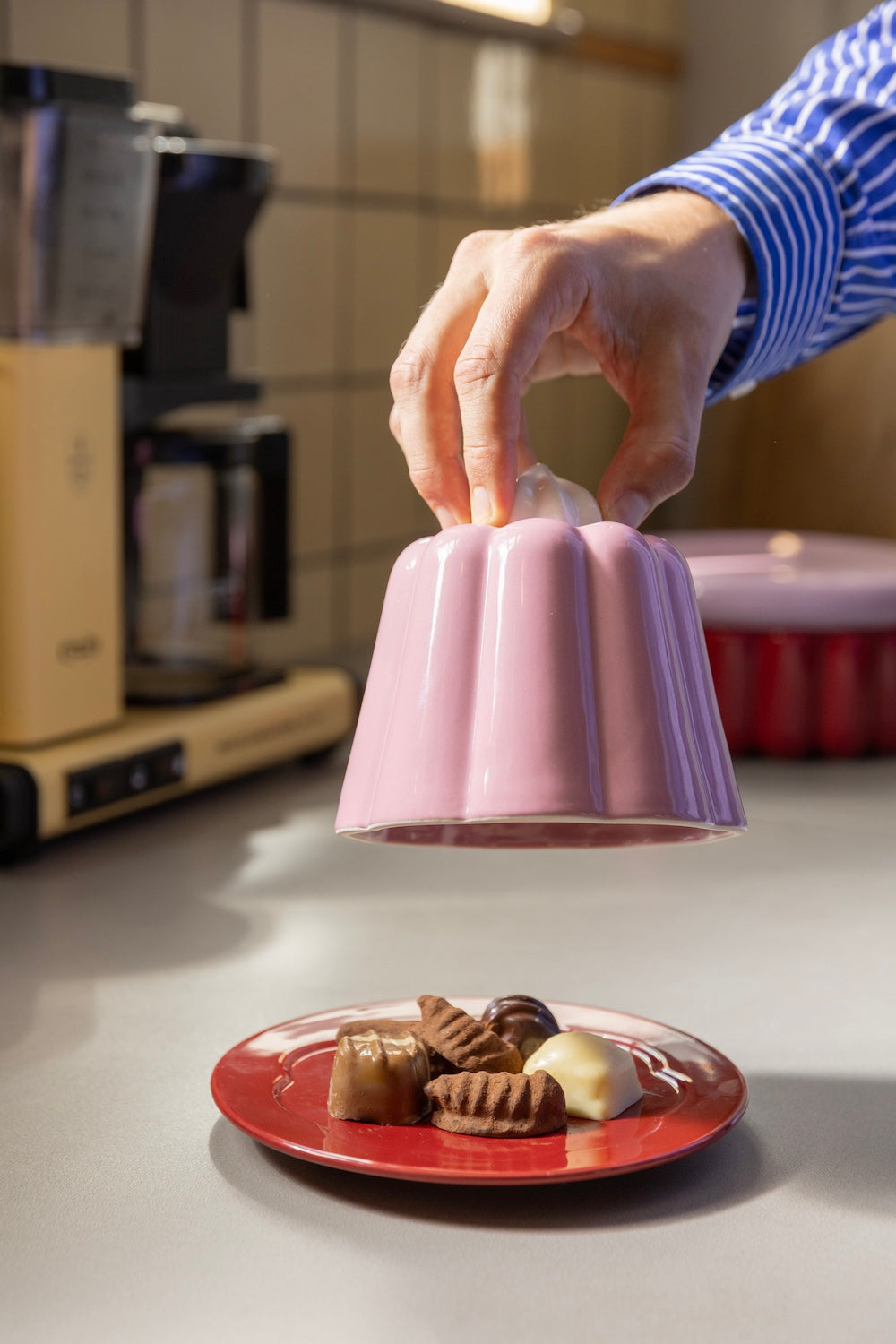 Hand lifting the pink pudding cloche to reveal biscuits on the red plate